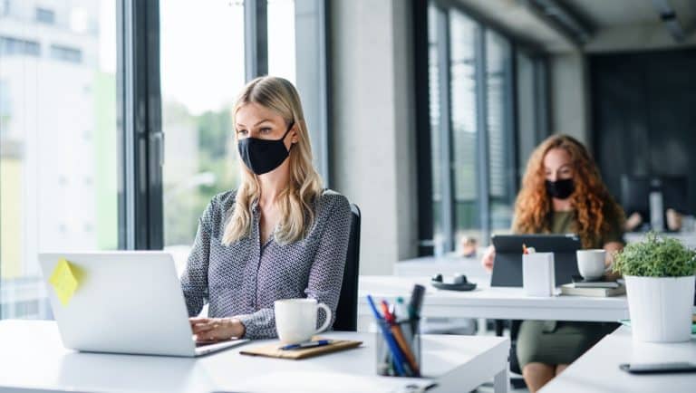 Duas moças sentadas em frente ao notebook no ambiente de trabalho. Elas utilizam máscaras de proteção anticovid.